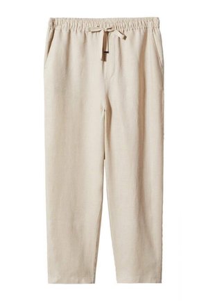 Personne portant un pantalon beige à cordon, des baskets blanches et une chemise blanche partiellement ouverte sur un haut blanc, debout sur un sol neutre.