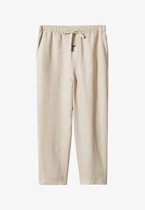 Personne portant un pantalon beige à cordon, des baskets blanches et une chemise blanche partiellement ouverte sur un haut blanc, debout sur un sol neutre.