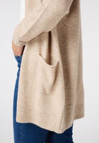Beige gebreide cardigan met geribde zoom en zijzak, met een zachte textuur en een relaxte pasvorm, gecombineerd met strakke blauwe jeans.