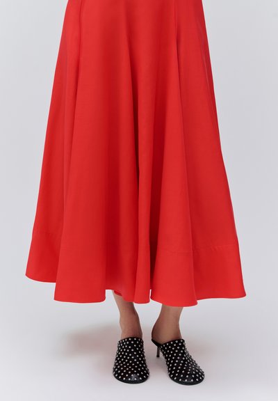 Jupe midi rouge fluide portée avec des mules à talons noires cloutées, sur fond blanc, montrant les jambes de la mi-mollet vers le bas.