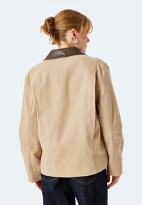 Beige jakke med en brun læderkrave, der har en afslappet pasform og synlige syninger. Bagfra ses et rent design og lange ærmer.