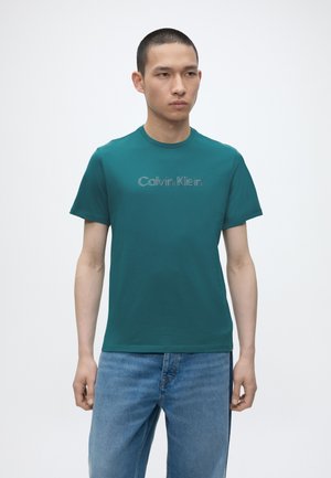 Νέος άνδρας που φοράει τιρκουάζ T-shirt Calvin Klein και μπλε τζιν, στέκεται μπροστά σε απλό φωτεινό φόντο, με ελαφρώς στραμμένο το σώμα προς τα δεξιά.