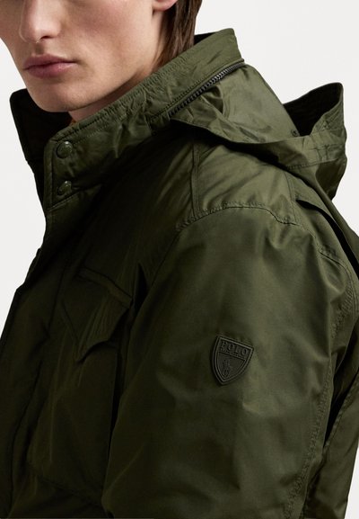 Olivgrüne Jacke mit Kapuze, ausgestattet mit einem Reißverschluss und Knopfakzenten. Beinhaltet ein Logopatch am oberen Arm und einen strukturierten Stoff.