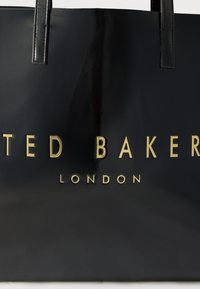 Črna bleščeča torbica z zlatim logotipom "Ted Baker London" in dvema črnima ročajema, vidnima na vrhu.