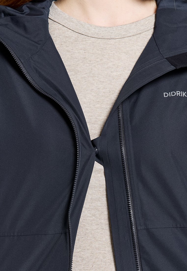 Schwarze Jacke mit Reißverschluss, teilweise geöffnet, über einem beigen gerippten Hemd, mit sichtbarem Markennamen "Didrik" auf der rechten Brustseite.