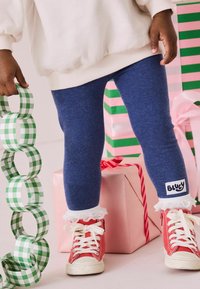 Granatowe legginsy z białą koronką, połączone z czerwonymi sneakersami. Dziecko trzyma zielone i białe w kratkę papierowe obrączki, a w tle widoczne są zapakowane prezenty.