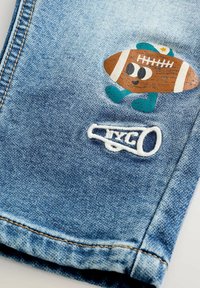 Tessuto di denim con un megafono bianco ricamato e dettagli di un colorato cartone animato a forma di pallone da calcio con scarpe e cappello sui jeans blu.