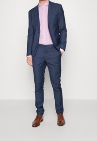 Mørkeblå habit med en tætsiddende pasform, struktureret stof og slim-fit bukser, kombineret med en lyserød skjorte og brune lædersko.
