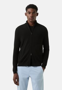 Černý svetr na zip vyrobený z měkkého materiálu, s vysokým límcem a bočními kapsami. Kombinován s světle modrými kalhotami.