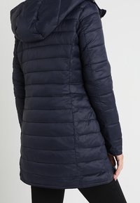 Mörkblå puffjacka med huva, med horisontellt quiltade sektioner, en slät, glänsande textur och en figurnära silhuett vid midjan.