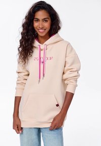 Beige mikina s kapucí a růžovými šňůrkami, s vyšitým textem. Obsahuje klokánkovou kapsu a žebrované manžety, vyrobená z měkké tkaniny.