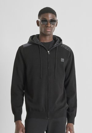 Zwarte rits hoodie met een gevoerde capuchon, voorzien van een logopatch, ribbelboorden en schouders met een glanzende afwerking. Zonnebril maakt de look af.