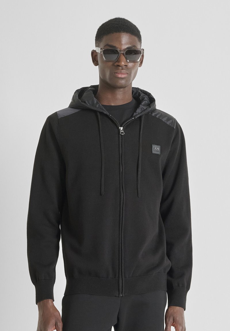 Felpa nera con zip e cappuccio foderato, con un patch con logo, polsini a costine e spalle con finitura lucida. Gli occhiali da sole completano il look.