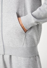 Lys grå zip-up sweatshirt lavet af blød stof, med ribbet manchetter og kant, samt sidelommer på hver side.