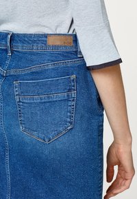 Falda de mezclilla en azul medio con un bolsillo trasero y costuras visibles. Etiqueta de la marca en marrón en la cintura. Textura suave y corte ajustado.