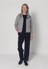 Veste en mélange de laine grise avec col montant et fermeture éclair, associée à un pantalon noir à jambes larges. Texture lisse, coupe moderne, et accessoires minimaux.