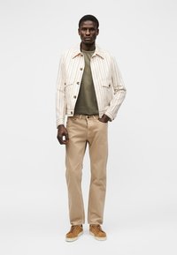 Άνδρας που φοράει μπεζ ριγέ σακάκι, πουκάμισο ελιάς, μπεζ παντελόνι και καφέ αθλητικά, στέκεται με το ένα χέρι στην τσέπη σε λευκό φόντο.