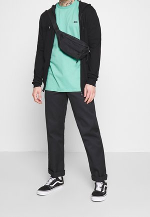 Schwarze Kapuzenjacke mit Reißverschluss, mintgrünes T-Shirt, schwarze Hose, schwarze Bauchtasche und schwarz-weiße Turnschuhe mit gestreiften Akzenten.