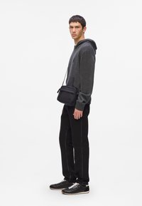 Giovane uomo che indossa un felpa grigia scura, pantaloni neri, sneakers nere e porta una piccola borsa a tracolla nera, in piedi di lato su uno sfondo bianco.
