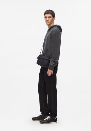 Jeune homme portant un sweat à capuche gris foncé, un pantalon noir, des baskets noires et portant un petit sac à bandoulière noir, se tenant de profil sur fond blanc.