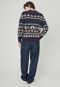 Pull en maille bleu marine avec des motifs festifs, comprenant des mugs de bière et des losanges, associé à un jean fluide bleu foncé et des baskets blanches.