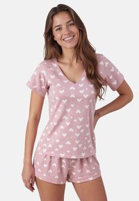 Set di pigiama in cotone rosa composto da una T-shirt con scollatura a V e pantaloni shorts. Il tessuto presenta un motivo ripetuto di cuori bianchi e una finitura leggermente testurizzata.