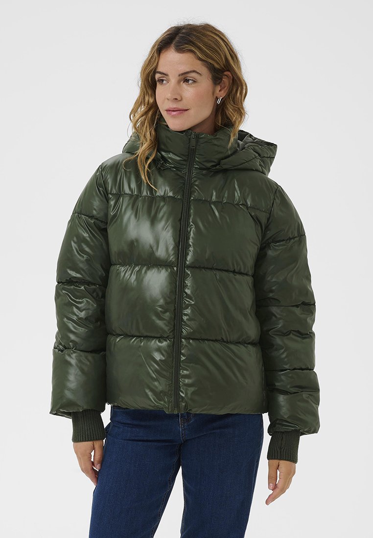 Grüne Pufferjacke mit glänzender Außenseite, horizontalen Nähten, Kapuze und gerippten Bündchen, kombiniert mit dunkelblauen Jeans.