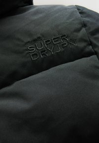 Čierna puffová bunda z látky so šitým logom "SUPER DRYJPN" čiernou niťou; hladká textúra s miernym leskom a vypchatým vzhľadom.
