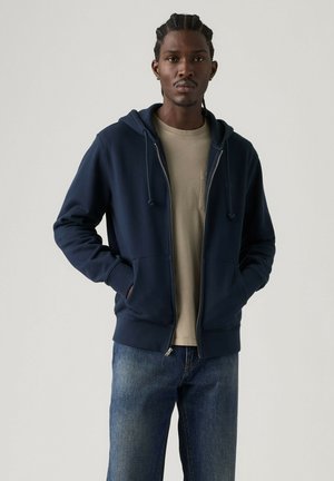 Mann mit geflochtenem Haar, der einen marineblauen Kapuzenpullover mit Reißverschluss über einem beigen Hemd und blauen Jeans trägt, mit den Händen in den Taschen vor einem einfarbigen Hintergrund steht.