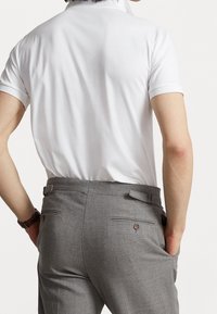 Man som bär en vit kortärmad polo skjorta instopad i grå kostymbyxor, med händerna i fickorna, sedd bakifrån mot en neutral bakgrund.