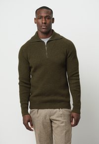 Olivgrüner, gerippter Strickpullover mit einem Viertelreißverschluss und einem V-förmigen Ausschnitt. Lange Ärmel mit schmalen Bündchen. Kombiniert mit hellfarbener Hose.