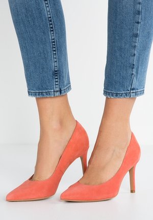 Décolleté in suede corallo con tacco alto e punta affusolata, caratterizzati da una texture liscia; indossati con jeans blue a culotte, che mettono in evidenza il tacco e la forma.