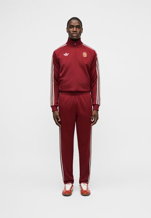 Homme debout, fait face à l'avant, portant un survêtement Adidas bordeaux avec des bandes blanches et des baskets rouges, sur un fond blanc uni.