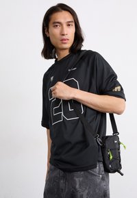 Black mesh jersey with white number "20" and logo. Paired with a small black crossbody bag featuring green accents and adjustable strap.