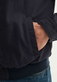 Veste marine avec un ourlet et des poignets à côtes, dotée d'une poche latérale et d'un tissu texturé. Gros plan de la main dans la poche, mettant en valeur les détails du design.