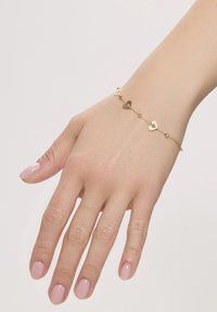 Złota bransoletka z małymi zaczepami w kształcie serca i okrągłymi koralikami, spoczywająca na dłoni z jasnoróżowym manicure. Gładka, błyszcząca tekstura.