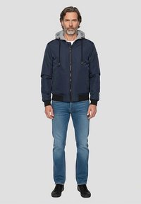 Marineblaue Bomberjacke mit grauer Kapuze, Reißverschluss, elastischen Bündchen und Saum. Kombiniert mit hellblauen Jeans und schwarzen Schuhen.