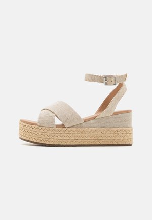 Sandale compensée en tissu beige avec des brides croisées à l'avant, bride de cheville avec boucle métallique, et semelle plateforme en fibres naturelles tressées.