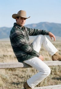 Homme avec un chapeau de cow-boy et une veste à carreaux assis sur une barrière en bois dans un champ, avec des montagnes en arrière-plan, regardant l'appareil photo.