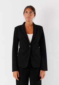 Blazer nero in tessuto strutturato con colletto a scaldacuore, chiusura con un bottone e due tasche frontali. Indossato sopra un top bianco.