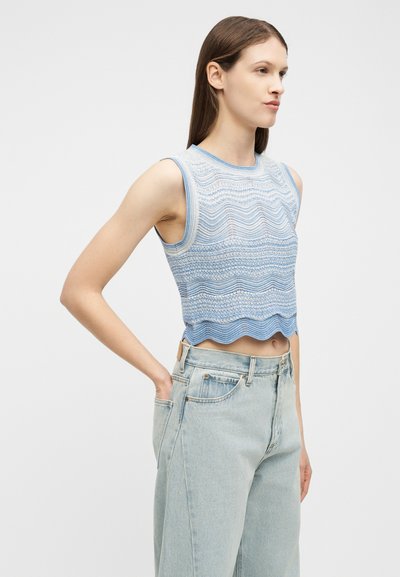 Νεαρή γυναίκα που φοράει αμάνικη, με σχέδιο, ανοιχτό μπλε κοντή μπλούζα και τζιν ψηλόμεσο με ανοιχτό πλύσιμο, στέκεται με το ένα χέρι στην τσέπη.