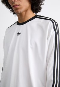 Balts garās piedurknes krekls ar melniem akcentiem. Ar trim melnām svītrām uz katra pleca un melnu Adidas logotipu uz krūtīm. Gluda auduma tekstūra.