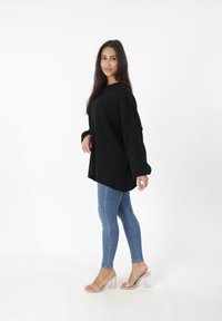 Maglione nero oversize a maglia con spalle scese, abbinato a jeans skinny blu chiaro e sandali con tacco a blocco trasparente, su sfondo bianco.