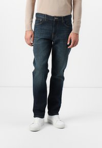 Moški v temno modrih kavbojkah, bež majici z dolgimi rokavi in belih športnih copatih, stoji na enostavni beli podlagi.