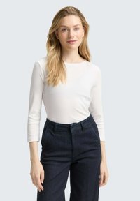 Weißes Langarmoberteil mit rundem Ausschnitt, kombiniert mit dunkelblauen High-Waist-Jeans. Glatte Textur, figurschnittiger Schnitt und subtile Nähdetails.
