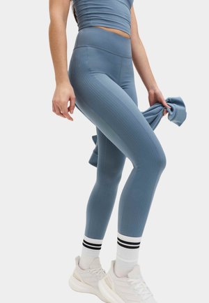 Kvinde iført blå højttaljede leggings, matchende crop top, hvide sneakers og hvide sokker med sorte striber, der holder en blå sweatshirt.