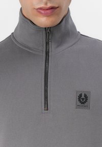 Pullover grigio con colletto alto e zip a metà. Caratterizzato da un tessuto testurizzato, una toppa nera con logo sul petto e dettagli di cucitura visibili.