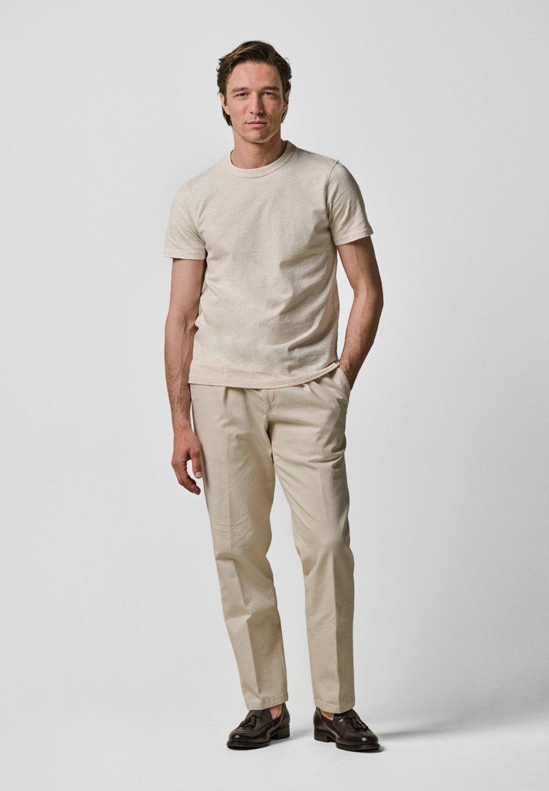 Beige t-shirt met korte mouwen, crèmekleurige beige broek, donkerbruine instap-schoenen, staande tegen een neutrale achtergrond, die eenvoudige, strakke lijnen en texturen benadrukt.