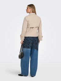 Vrouw draagt een beige korte jas en wijde blauwe spijkerbroek met zwart bloemenpatroon, houdt een zwarte handtas vast, staande met het gezicht naar links.