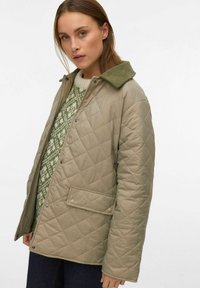 Veste beige matelassée avec col en velours côtelé vert, boutons-pression et une poche avant, portée par-dessus un pull vert et blanc à motifs.
